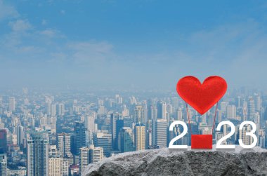 Kırmızı Kumaş Kalp 2023 beyaz metinli balonu sever. Modern şehir kulesi ve gökdelenin tepesindeki kaya dağında. Mutlu yıllar ve Sevgililer Günü 2023 konsepti.