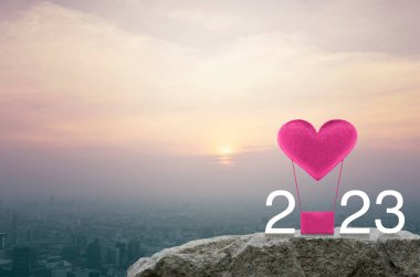Pembe kumaşlı kalp, 2023 beyaz metinli hava balonuna bayılıyor. Gün batımında şehir manzarası, klasik tarz, mutlu yıllar ve 2023 Sevgililer Günü konsepti.
