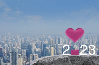 Pembe kumaşlı kalp, 2023 beyaz metinli hava balonunu modern şehir kulesi ve gökdelen üzerindeki kaya dağında seviyor. Sevgililer Günü konsepti.