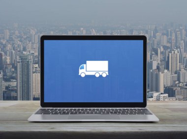 Delivery truck flat icon on modern laptop computer screen on wooden table over city tower and skyscraper, Business transportation online service concept