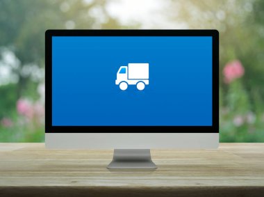 Masaüstü modern bilgisayar monitöründeki kargo kamyonu düz simgesi parktaki bulanık pembe çiçek ve ağacın üzerindeki ahşap masa üzerinde, iş ulaşımı çevrimiçi servis konsepti