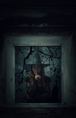 Cadılar Bayramı cadısı sessiz işareti gösteriyor. Dudak üstünde parmak izi olan eski hasarlı ahşap pencerenin üzerinde duruyor. Duvarları haç, kilise, kuşlar, ölü ağaç ve ürkütücü bulutlu gökyüzü, Cadılar Bayramı gizemi.