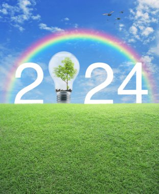 Mavi gökyüzü, beyaz bulutlar ve gökkuşağı üzerinde yeşil çimlerin üzerinde ağaç olan 2024 beyaz metin ve ampul 2024 ekolojik kapak konsepti mutlu yıllar.