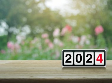 Ahşap masa üzerinde 2024 harfli ters saat parkta bulanık pembe çiçek ve ağaç üzerinde mutlu yıllar 2024 kapak konsepti