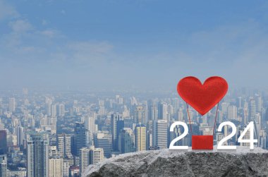 Kırmızı Kumaş Kalp, 2024 beyaz yazılı balonu sever. Modern şehir kulesi ve gökdelenin üzerindeki kaya dağında. Mutlu yıllar ve Sevgililer Günü 2024 konsepti.