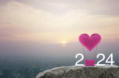 Pembe kumaşlı kalp, 2024 beyaz metinli hava balonuna bayılıyor. Gün batımında şehir manzarası, klasik tarz, mutlu yıllar ve 2024 Sevgililer Günü konsepti.