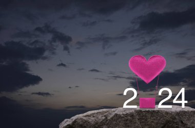 Pembe kumaşlı kalp, 2024 beyaz metinli hava balonuna bayılıyor. Gün batımında gökyüzünde kaya üzerinde. Mutlu yıllar ve 2024 Sevgililer Günü kavramı.