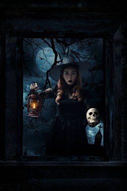 Cadılar Bayramı cadısı antik lamba ve kafatasını eski hasarlı ahşap pencerenin önünde duruyor. Duvarları haç, kilise, kuşlar, ölü ağaç ve ürkütücü bulutlu gökyüzü, Cadılar Bayramı gizemi.