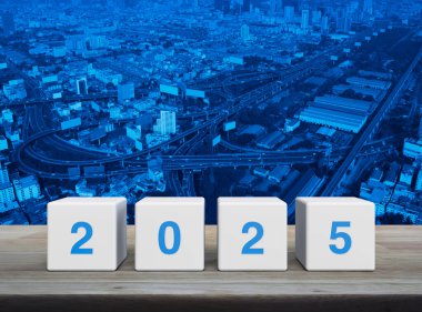 Modern şehir kulesi, sokak, ekspres yol ve gökdelen üzerindeki ahşap masa üzerinde 2025 harfli beyaz blok küpleri. Mutlu yıllar 2025 kapak konsepti.