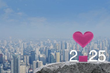 Pembe kumaşlı kalp, 2025 beyaz yazılı balonu modern şehir kulesi ve gökdelenin üzerindeki kaya dağında seviyor. Mutlu yıllar ve Sevgililer Günü 2025 konsepti.