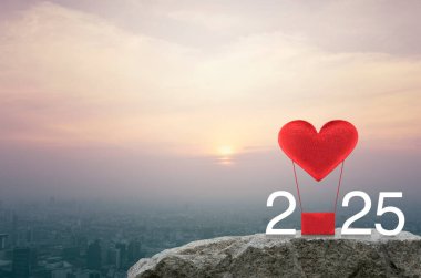 Kırmızı Kumaş Kalp, 2025 beyaz metinli hava balonuna bayılıyor. Gün batımında şehir manzarasının havadan görünüşü, klasik tarz, yeni yıl ve 2025 Sevgililer Günü konsepti.