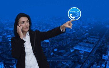 İş kadını cep telefonuyla konuşuyor ve modern şehir kulesi, sokak, ekspres yol ve gökdelen üzerinde telefon ve e-posta ikonu düğmesini işaret ediyor, bizimle ve müşteri hizmetleri konseptiyle irtibata geçiyor.