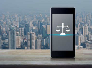 Şehir kulesi ve gökdelen üzerindeki ahşap masanın üzerinde modern akıllı cep telefonu ekranında yasalara uygun simge, iş hukuku hizmeti online konsepti