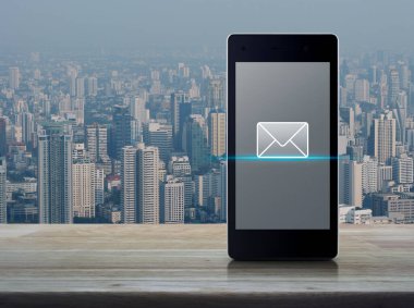 Şehir kulesi ve gökdelen üzerindeki ahşap masada modern akıllı cep telefonu ekranında e-posta düz simgesi, iş iletişimi çevrimiçi konsepti