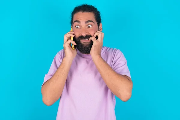 恐ろしいおかしなコーカサス人男性は 電話を保持し 爪を噛む青い背景の上にヴァイオレットTシャツを身に着けています — ストック写真