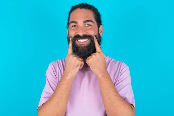 ハッピーコーカサス人男性は 歯痛のある笑顔で青い背景にヴァイオレットTシャツを着用し 口の近くで指を指し 陽気な笑顔を強制する — ストック写真
