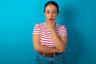 Çizgili tişört giyen düşünceli bir kadın elini çenesinin altında tutuyor, doğrudan kameraya bakıyor, ilgi çekici bir şey dinliyor. Gençlik kavramı.