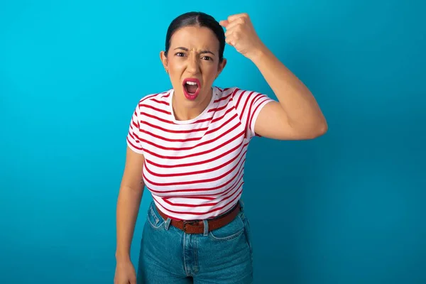 Fierce woman wearing striped T-shirt holding fist in front as if is ready for fight or challenge, screaming and having aggressive expression on face.
