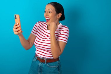 Çizgili tişört giyen mutlu bir kadın e-posta ile iyi haberler alıyor ve ayakta durup cep telefonuna bakarken başarıyı kutluyor..