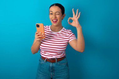 Çizgili tişört giyen mutlu bir kadın akıllı telefonuna mesaj yolluyor ya da selfie çekiyor ve eliyle imzasını atıyor..