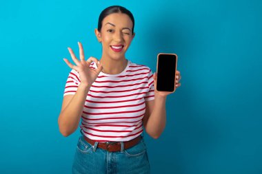 Çizgili tişört giyen heyecanlı bir kadın akıllı telefon boş ekranı gösteriyor, göz kırpıyor ve el işareti yapıyor. Reklam kavramı.