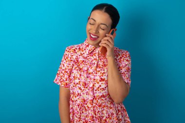 Çiçekli elbise giyen komik kadın mutlu bir şekilde gülüyor, telefonla konuşuyor, arkadaşı tarafından eğlendiriliyor, gözlerini kapatıyor..