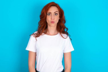 Kızıl saçlı kadın mavi arka plan üzerine beyaz tişört giyiyor. Tiksinti, isteksizlik, gergin bakışlar, somurtkan bir yüz ifadesi var..
