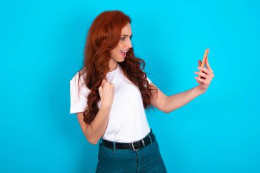 Beyaz tişört giyen neşeli kızıl kadın e-posta ile iyi haberler alıyor ve ayakta durup cep telefonuna bakarken başarıyı kutluyor..