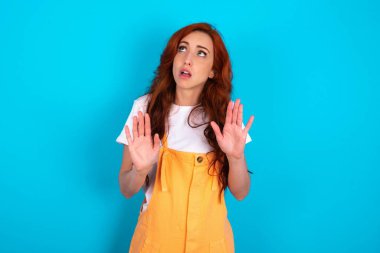 Mavi arka planda turuncu giyen kızıl saçlı kadın avuçlarını öne doğru uzatıyor ve tavandan korkuyla bakıyor kendini görünmez tehlikeden korumaya çalışıyor. Ağzını açıyor..