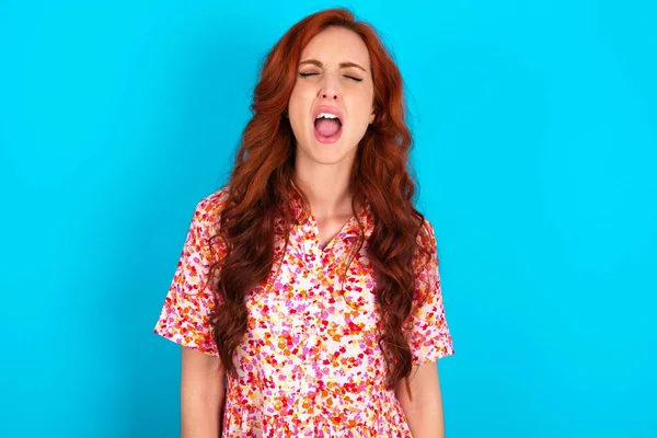 Stressful redhead woman wearing floral dress over blue background screams in panic, closes eyes in terror, keeps hands on head, finds out terrified news, can\'t believe it.