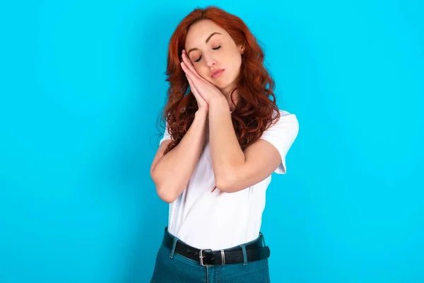 Relax and sleep time. Tired redhead woman wearing white T-shirt over blue background with closed eyes leaning on palms making sleeping gesture.