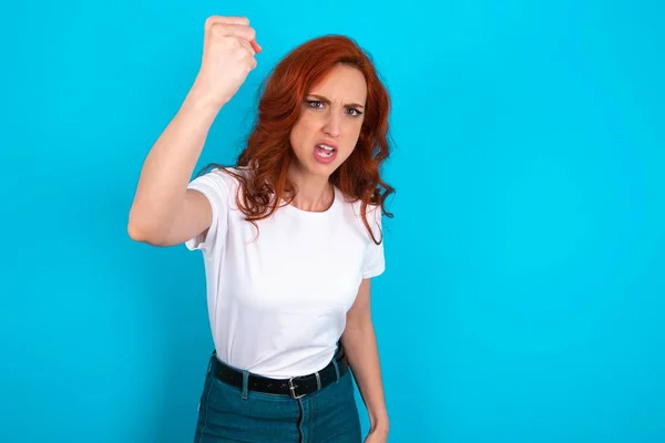 Fierce redhead woman wearing white T-shirt over blue background holding fist in front as if is ready for fight or challenge, screaming and having aggressive expression on face.