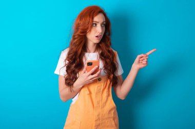 Afallamış kızıl saçlı kadın mavi arka planda turuncu giyiniyor. Yan yan duruyor. Sağ fotokopi odası, ürün öneriyor, şaşırtıcı şeyler görüyor.