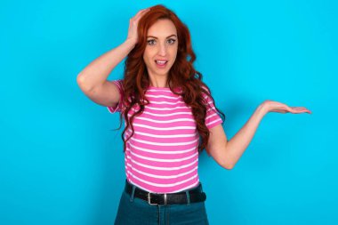 Şok oldum, şaşkın kızıl saçlı kadın pembe çizgili tişört giyip mavi arka planda el ele tutuşmayı teklif ediyor.