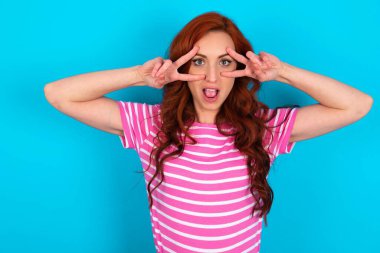 Mavi arka planda pembe çizgili tişört giyen neşeli, pozitif saçlı bir kadın, gözleri ve ağzı açık V işaretini gösteriyor.