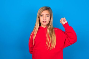 Güzel beyaz kız mavi arka planda kırmızı kazak giyiyor ciddi, güçlü ve asi hissediyor, yumruğunu kaldırıyor, protesto ediyor ya da devrim için savaşıyor.. 