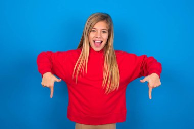 Mavi atölyenin arka planında kırmızı sweatshirt giyen beyaz bir kız. Pozitif bir ifadeyle iki işaret parmağıyla işaret ediyor. Geniş ilgi çekici bir gülümsemesi var. Şuraya bakın, lütfen..