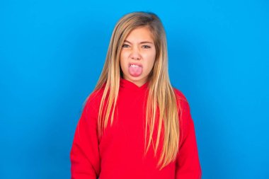 Body language. Disgusted stressed out caucasian teen girl wearing red sweatshirt over blue background, frowning face, demonstrating aversion to something clipart