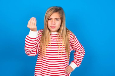 Shot of frustrated blonde teen girl wearing striped t-shirt over blue wall gesturing with raised hand doing Italian gesture, frowning, being displeased and confused with dumb question.