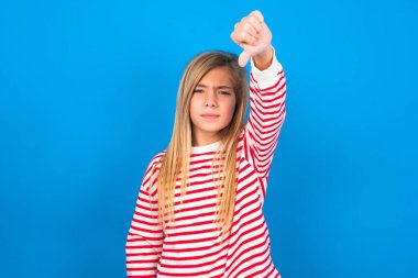 Mavi arka planda çizgili tişört giyen hoşnutsuz genç kız tasvip etmiyor, baş parmağını aşağıda tutuyor, hoşnutsuzluğunu ifade ediyor, hoşnutsuzluğunu ifade ediyor. Olumsuz duygular.