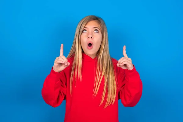 青い背景に赤いスウェットシャツを着ている美しい慎重な十代の少女は驚き 見て驚き 何か奇妙なものを示す指で指を指さす — ストック写真