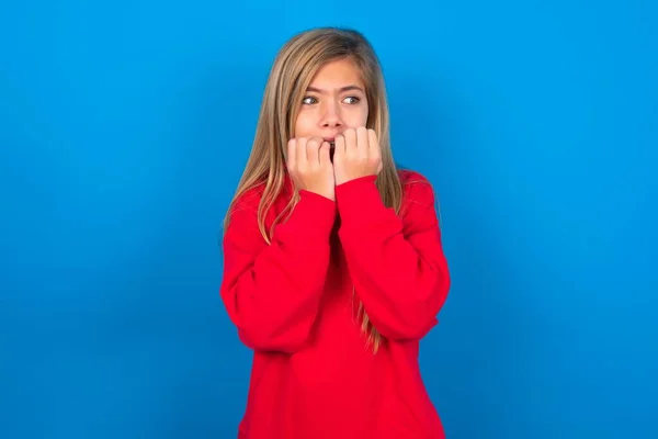 stock image Caucasian teen girl wearing red sweatshirt over blue studio background covering his mouth with hands scared from something or someone biting nails.