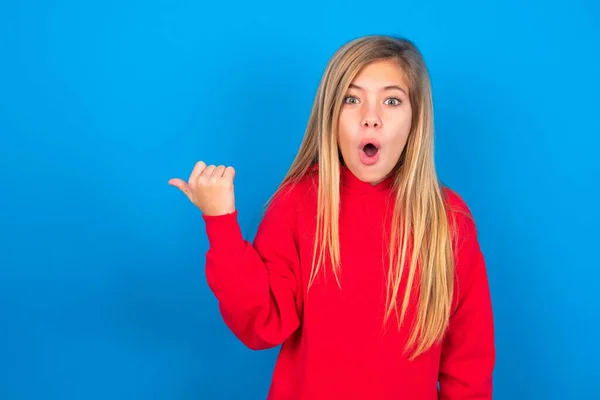 ショックを受けた白人の十代の少女は 親指で青い背景のポイントの上に赤いスウェットシャツを着て 何かを示しています これをチェック 広告コンセプト — ストック写真