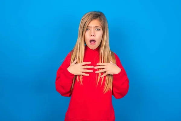 stock image Caucasian teen girl wearing red sweatshirt over blue studio background keeps hands on chest feeling shocked and scared, mouth widely opened, stares at camera  