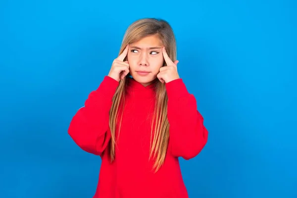 思慮深い表情で青いスタジオの背景に赤いスウェットシャツを着て 何かを考える白人少女 — ストック写真