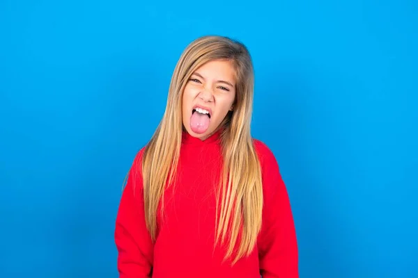 ブロンドの慎重な十代の少女は 幸せで面白い顔の笑顔と舌を示す青い背景に赤いスウェットシャツを着ています — ストック写真