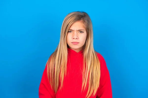 ブロンド慎重な十代の少女は青い背景に赤いスウェットシャツを着て眉毛が何かに不満を抱いている — ストック写真