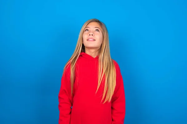 チャーミングな慎重な十代の少女は 彼女が奇妙なものを見ている青い背景の上に赤いスウェットシャツを着ています — ストック写真