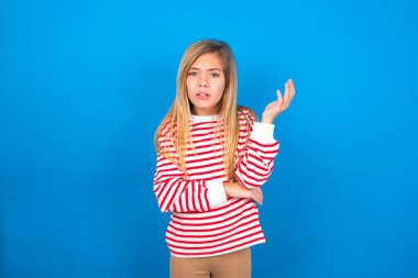 Studio shot of frustrated teen girl wearing striped shirt over blue background gesturing with raised palm, frowning, being displeased and confused with dumb question.