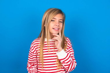 Kaygısız, başarılı genç kız mavi arka planda çizgili gömlek giyiyor çene hattına dokunuyor kameraya bakıyor başını yana eğmiş, beyaz dişlere bayılıyor. Diş sağlığı konsepti.